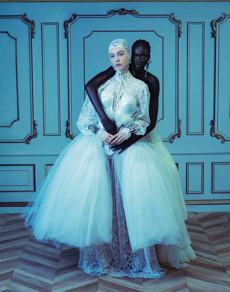 Two women in elaborate white gowns embrace against a blue paneled wall and herringbone floor, fashion portrait, studio lighting.