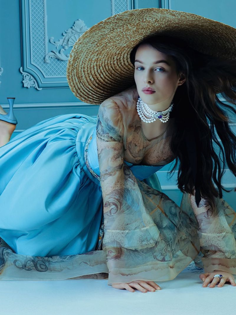 Woman wearing a wide-brimmed hat, pearl choker, and blue dress poses indoors with an ornate wall and flooring in a beauty fashion shot.