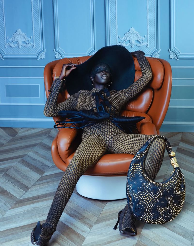 Fashion editorial shot of a woman in a patterned jumpsuit and wide-brimmed hat, sitting in a leather chair with a patterned handbag.