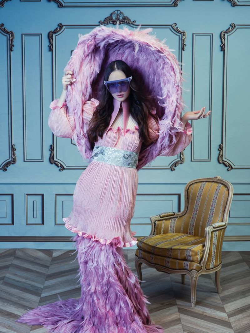 Adult female wearing an elaborate pink feathered hat and dress poses in front of a blue paneled wall with an ornate chair.