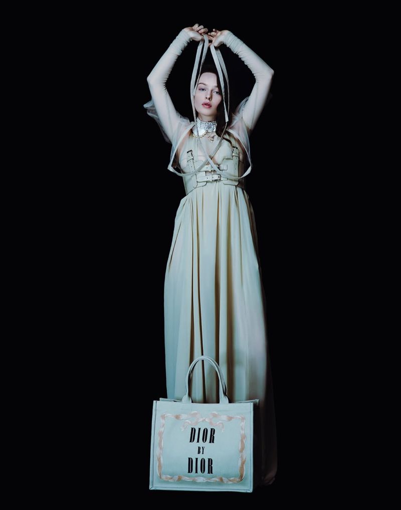 A model wearing an elegant dress and holding white straps, standing against a dark background, and presenting a Dior by Dior tote bag.