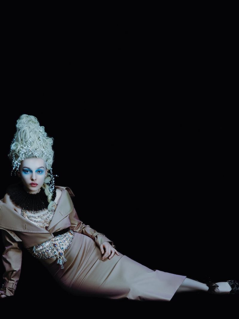 A woman with blue eyeshadow and a white wig lies down in a beige dress in a dark, high-fashion portrait with studio lighting.