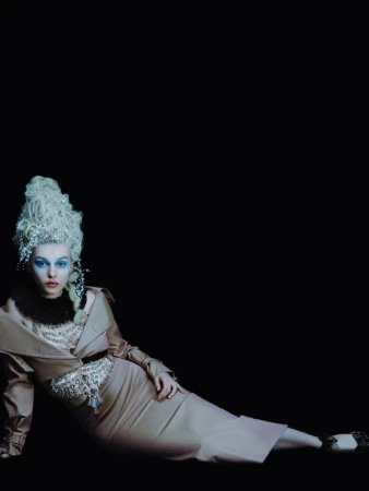 A woman with blue eyeshadow and a white wig lies down in a beige dress in a dark, high-fashion portrait with studio lighting.