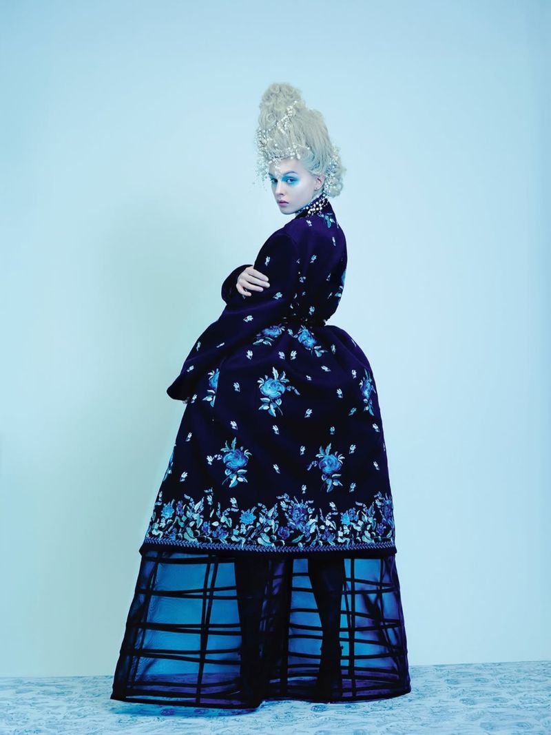 Woman in elaborate dark blue floral dress and tall blonde wig poses in a light blue photo studio with arms crossed and looking at the camera.