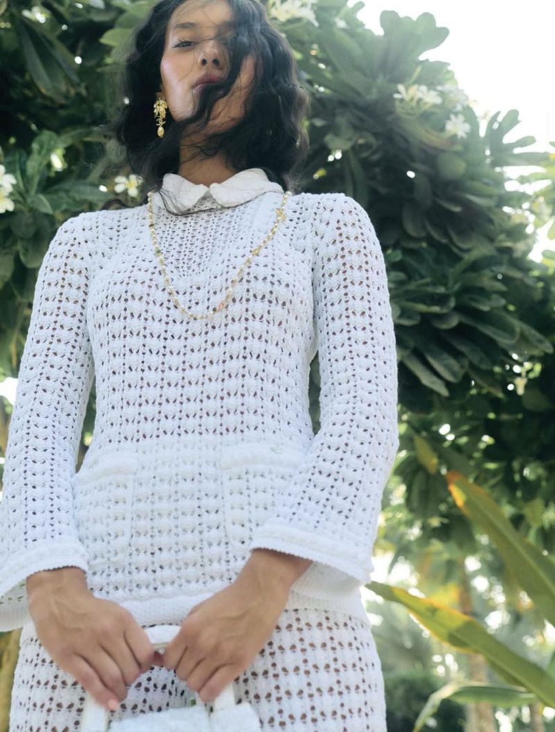 A woman poses in a crochet dress with a white collar and handbag, accented by gold jewelry, against a lush green backdrop with white flowers.