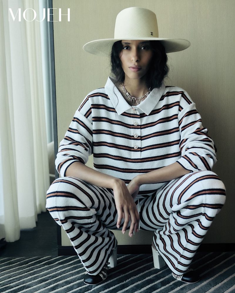 A woman in a striped outfit and hat squats indoors, illuminated by window light, showcasing modern fashion against a neutral backdrop.
