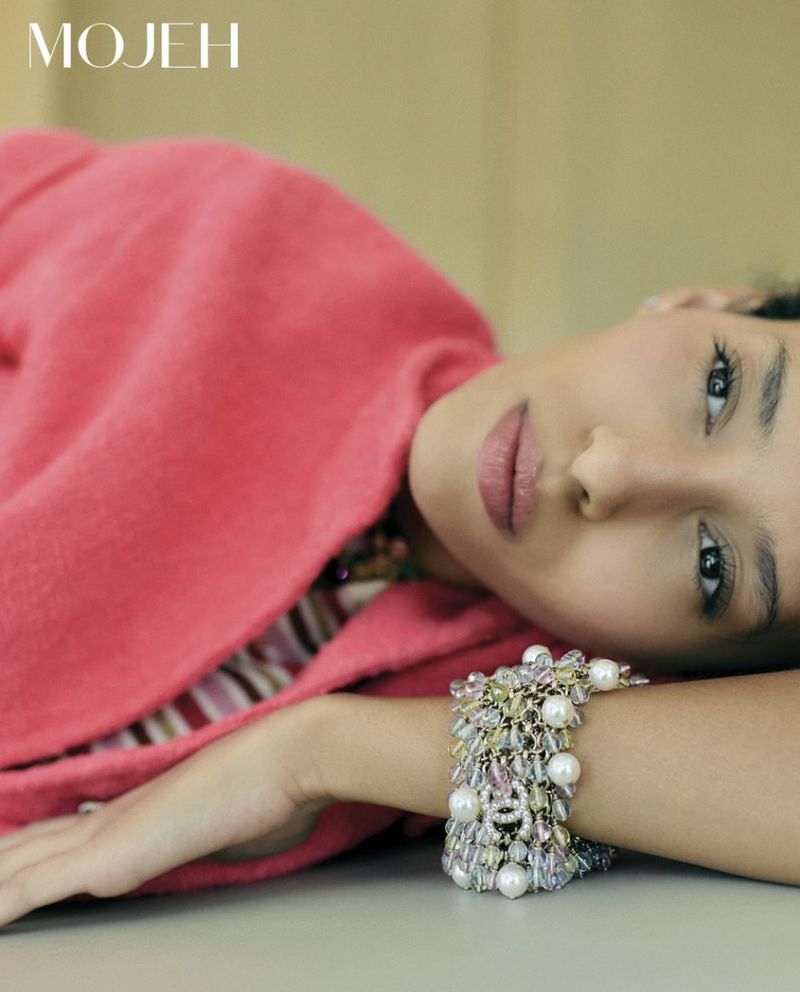 Woman lies with bejeweled bracelet, wearing a pink coat, captured in a studio shot with soft lighting for a fashion editorial.