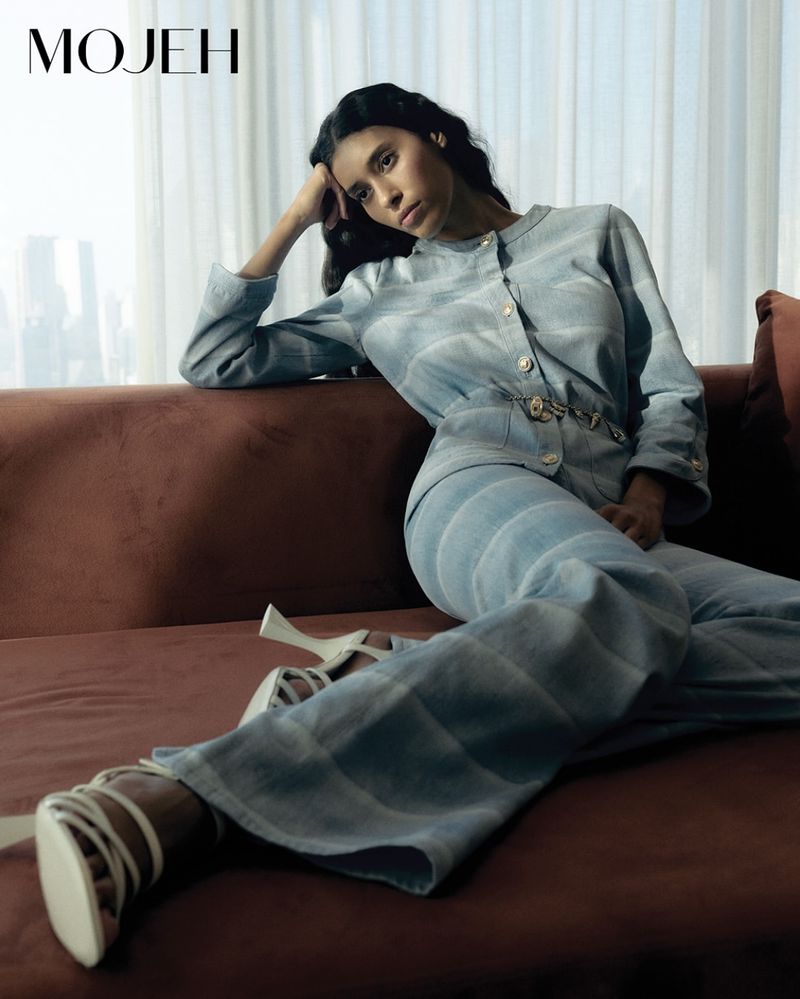 A woman in a blue striped pantsuit poses on a rust-colored couch with natural light streaming in from a window, showcasing fashion.
