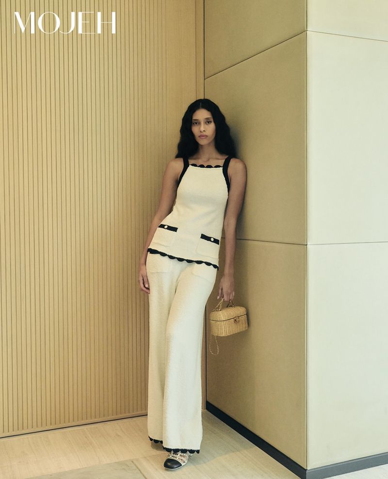 A woman in a cream-colored outfit poses against a modern wall, holding a purse, indoors, styled for a fashion editorial.