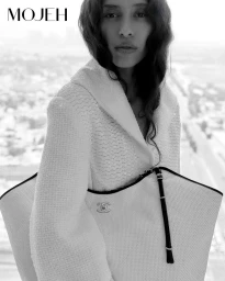 Black and white portrait of model Allana Neiva in a textured white jacket holding a large quilted Chanel tote bag by a window.