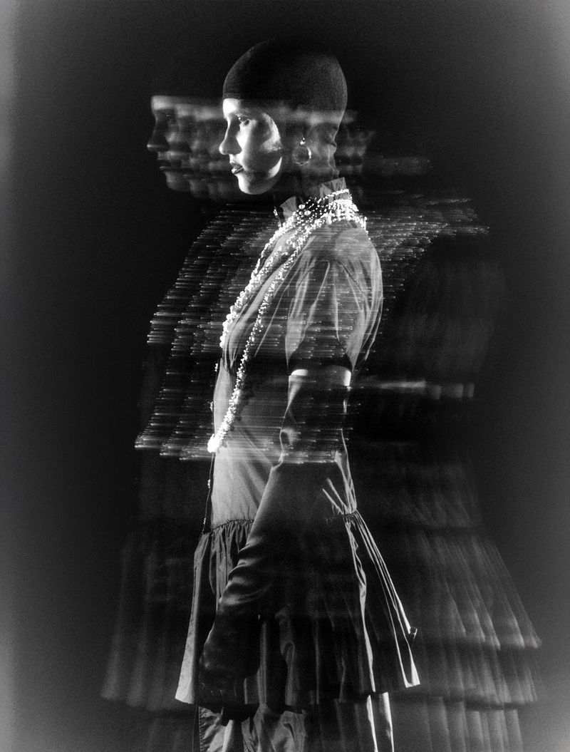 Black and white long exposure fashion shot of a woman wearing a dress, hat, and jewelry, with a blurred effect, side view.