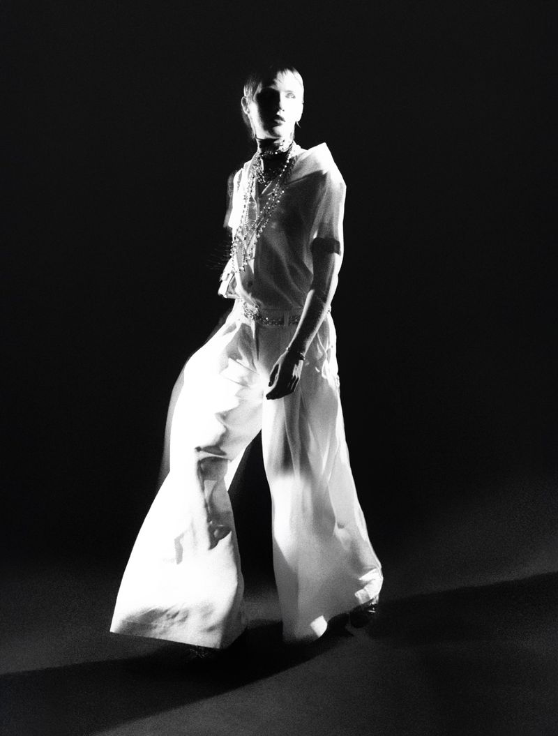 Model in white trousers and layered necklaces under dramatic low-key lighting against a black background