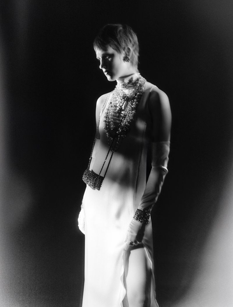 A woman stands in a dimly lit room, wearing an elegant white gown with layered beaded necklaces and wrist cuffs, accessorized with a small purse.