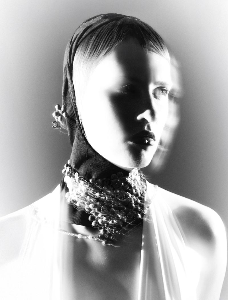 Monochrome portrait of a woman wearing a headscarf, pearl choker, and dress with motion blur, against a light gray background in studio lighting.