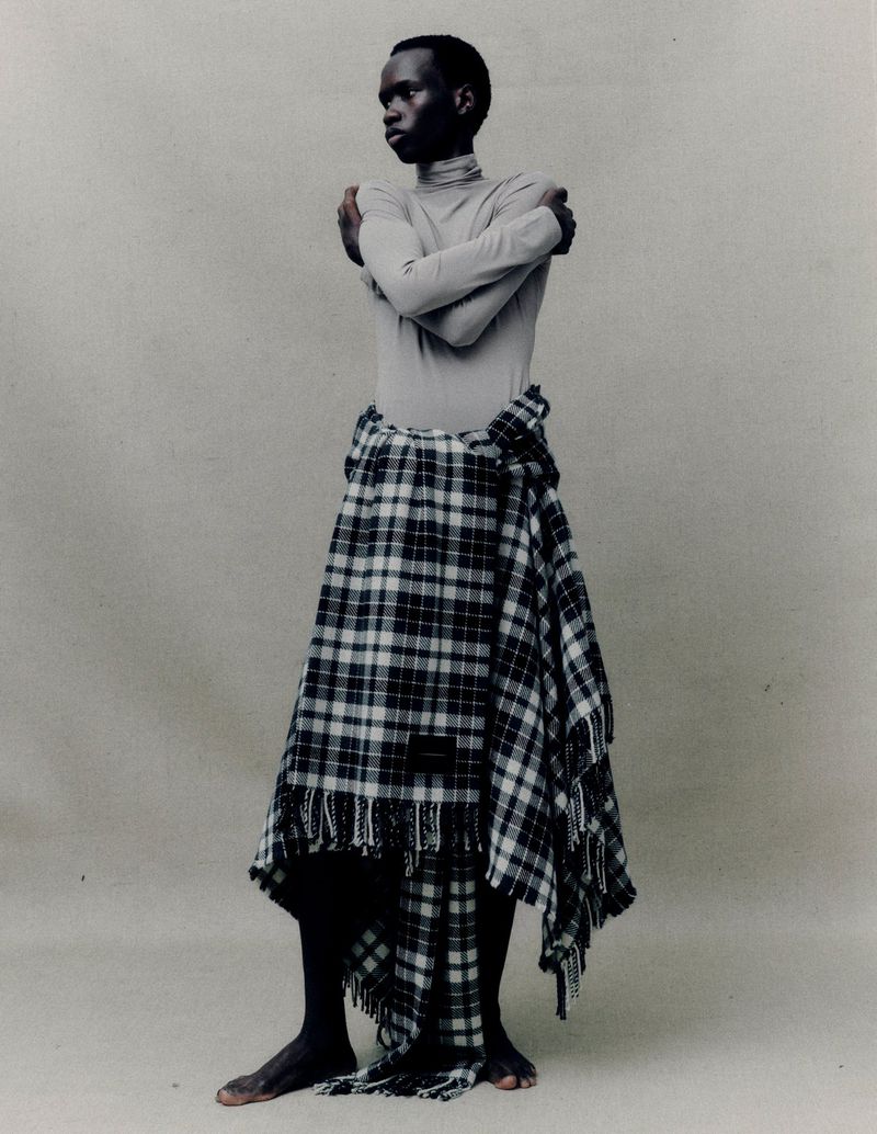 Full shot of a dark-skinned adult male, barefoot, in light-grey turtleneck and tartan wrap standing against a plain grey background in studio lighting.
