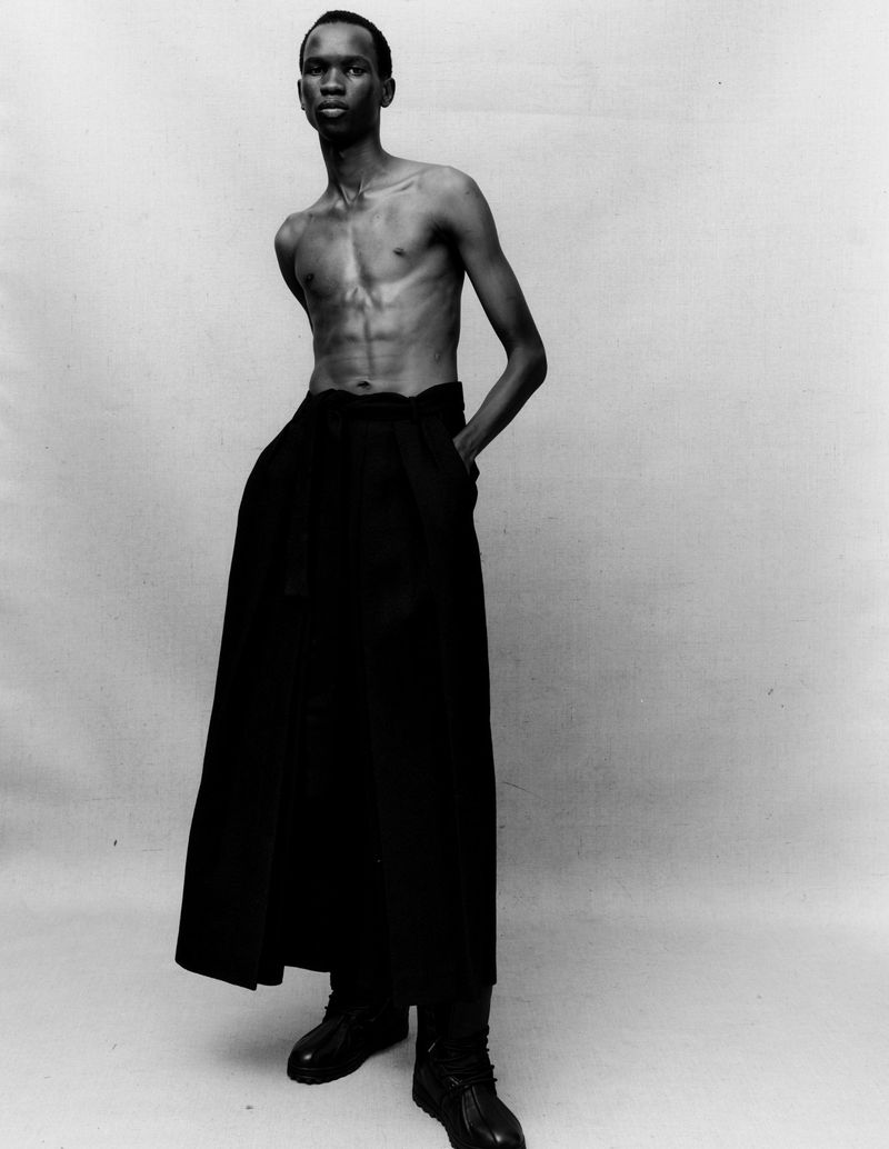 A topless African-American man stands in a studio wearing black, high-waisted pants and shoes against a neutral grey backdrop.