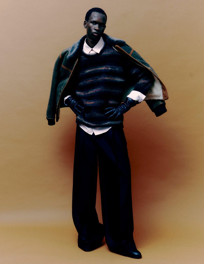 A dark-skinned man poses in black wide-leg pants and a striped sweater; an aviator jacket is draped over his shoulder against tan backdrop.