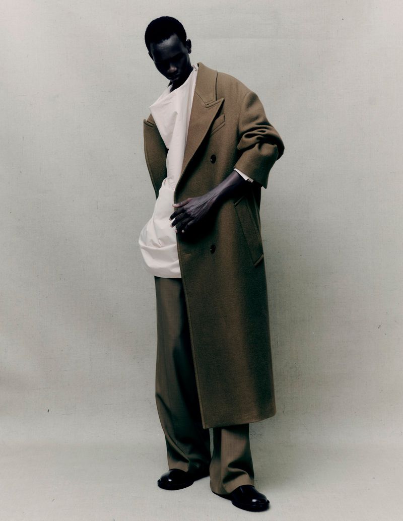 Male fashion model in studio poses in a full-length shot, wearing an olive overcoat, oversized white shirt, and wide-legged trousers.
