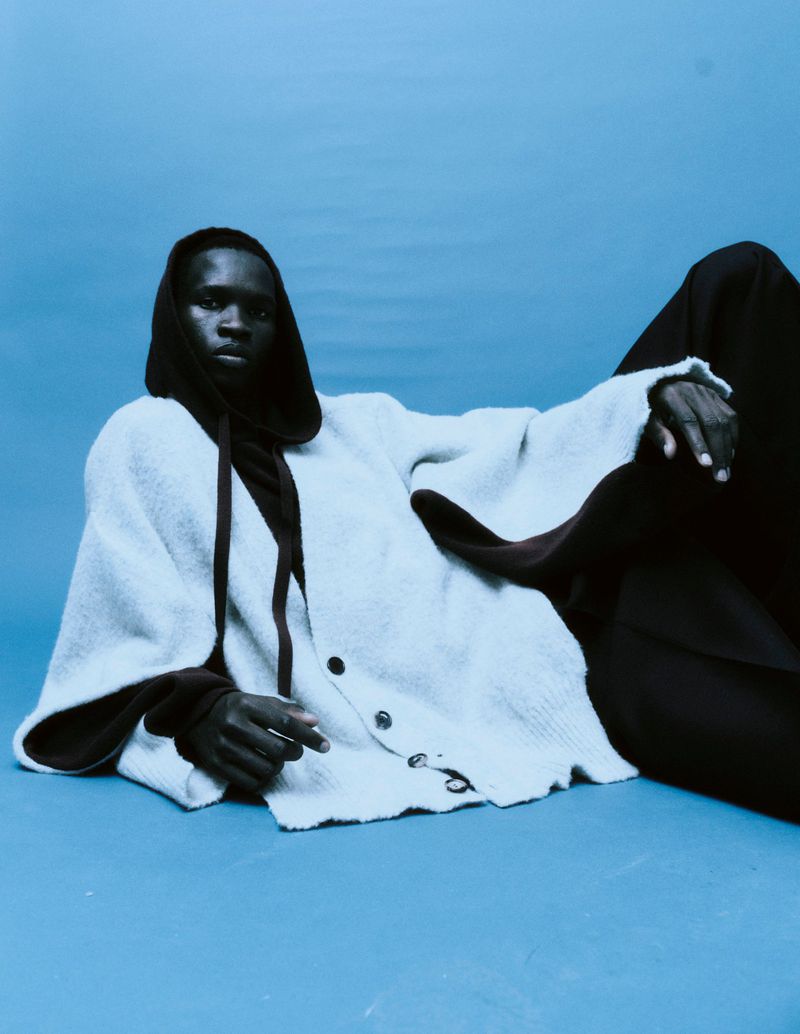 Dark-skinned model in hooded cardigan and pants poses in a studio setting, lying down on a blue backdrop in a fashion editorial style.