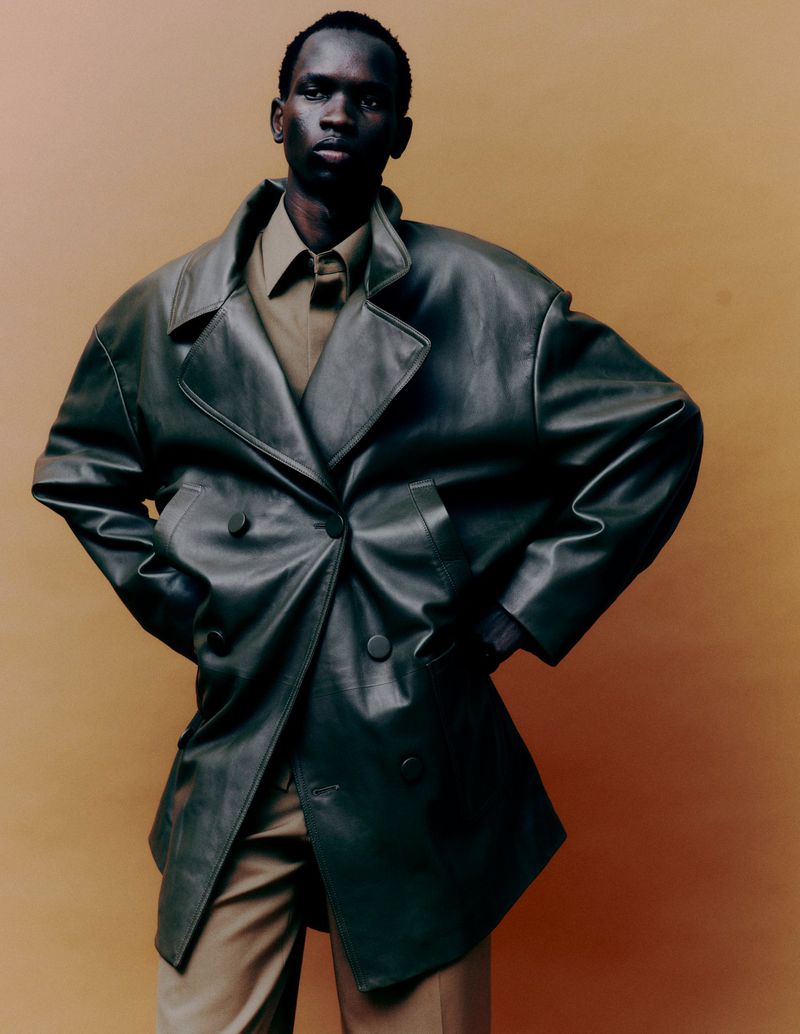 Dark-skinned man in a green leather double-breasted coat against a tan background. Stylish and confident studio fashion shot.