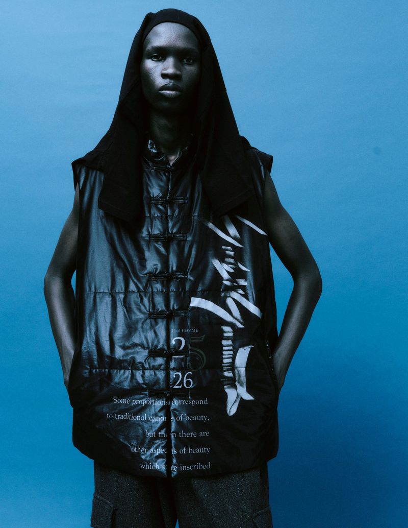 Adult male poses in a black hooded vest with text against a blue studio backdrop, presenting a fashion-forward editorial image.
