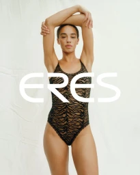 Model posing with arms raised overhead wearing a black patterned swimsuit against a pale fabric backdrop