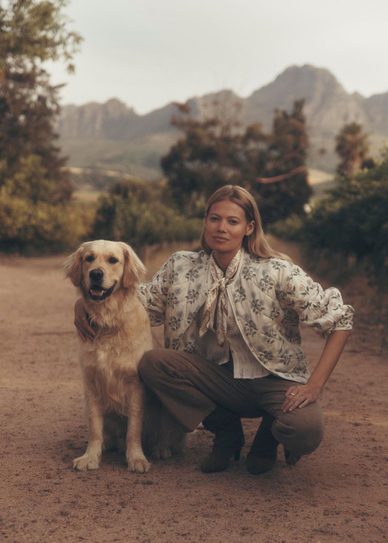 Blonde woman crouches with a golden retriever on a dirt road, with mountains and greenery in the background in an outdoor setting.