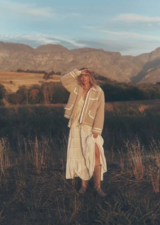 Woman in field wearing a long dress, beige jacket, and boots, shielding eyes with hand. Mountains in the background under a blue sky.
