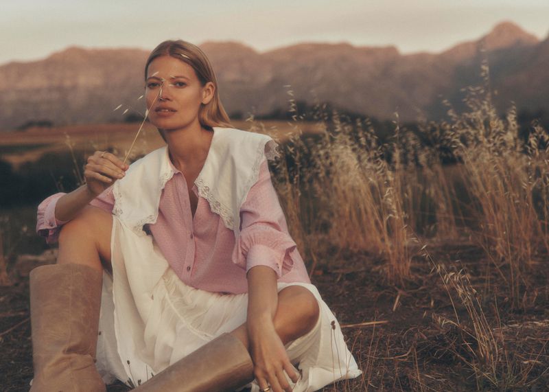 A blonde adult female model sits outdoors in a pink blouse and white skirt, holding wheat, set against a scenic mountain backdrop.