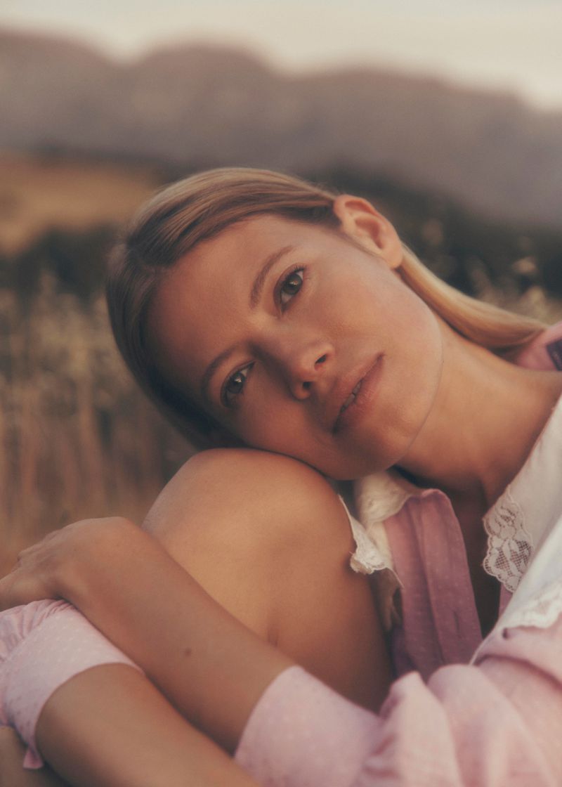 Portrait of a blonde adult female with arms around her legs outdoors in a natural environment with mountains in the background.