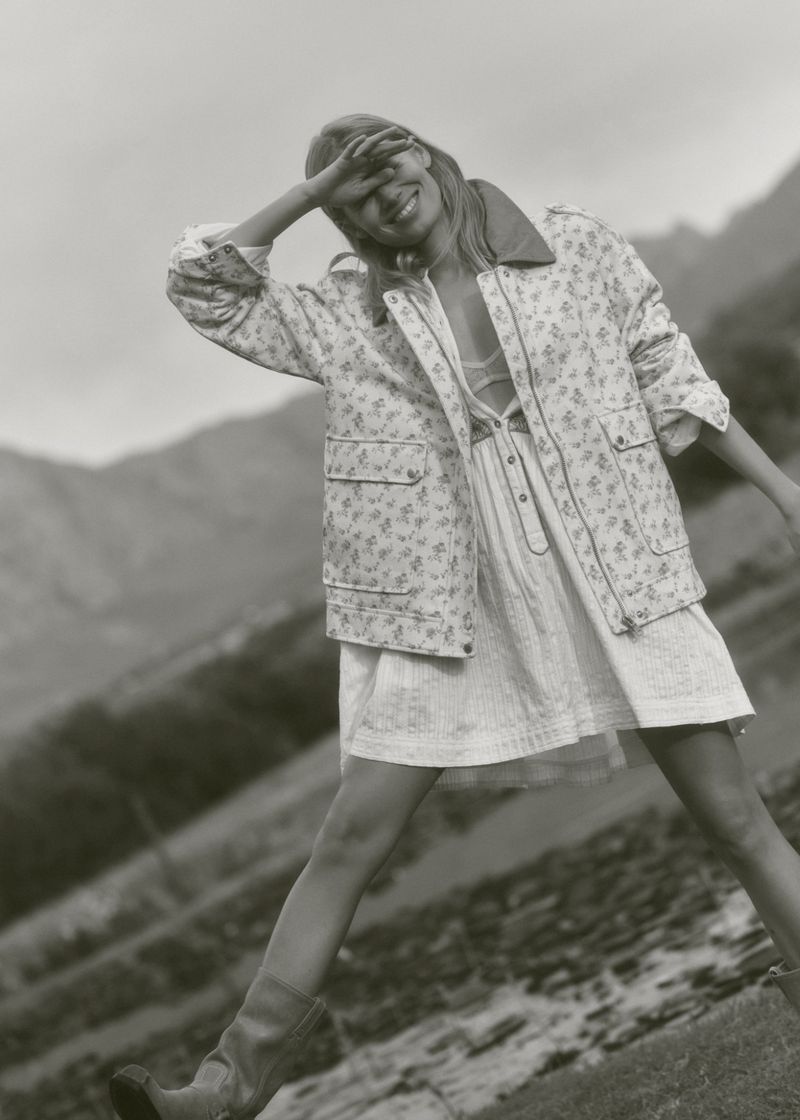 A woman in a floral jacket and dress stands outside with mountains in the background, shielding her eyes from the sun.
