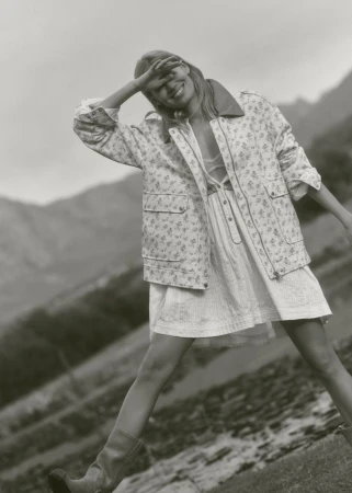 A woman in a floral jacket and dress stands outside with mountains in the background, shielding her eyes from the sun.