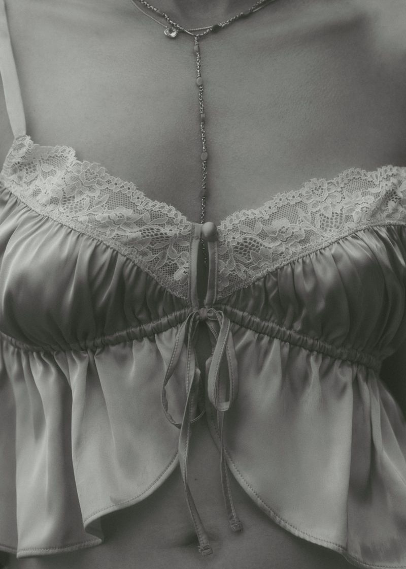 Close-up of a woman's torso wearing a camisole top with lace trim and a long, delicate silver necklace against a gray background.