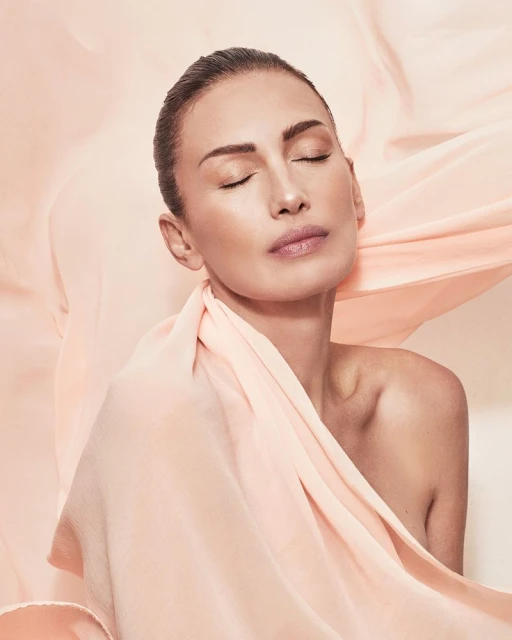 Close-up portrait of a woman with her eyes closed, draped in a peach-colored fabric, against a peach-toned studio background.
