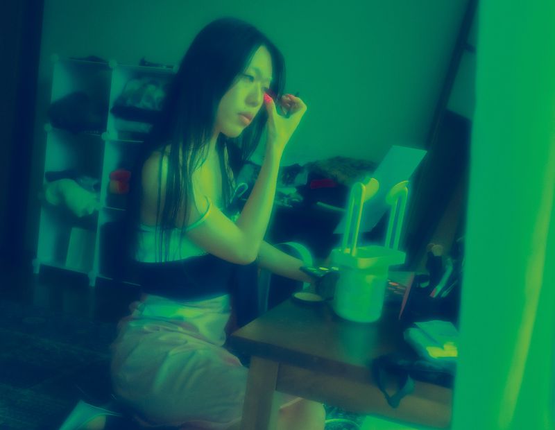 Woman applies makeup at vanity with beauty mirror in an indoor room, bathed in a green hue, creating an atmospheric beauty setting.