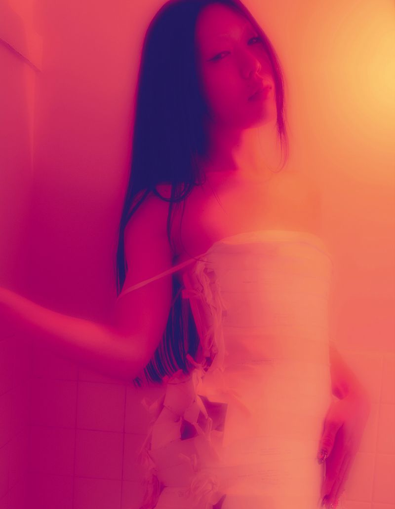 A woman with long dark hair poses in what appears to be a towel or wrap, bathed in red and orange lighting in an indoor setting.
