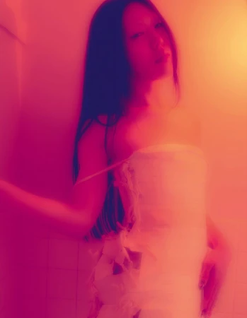 A woman with long dark hair poses in what appears to be a towel or wrap, bathed in red and orange lighting in an indoor setting.