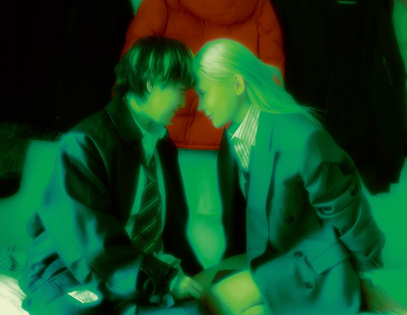 Two people face each other in green-toned photo. An adult male in a suit and tie looks at an adult female in a blazer, indoors, near clothes.