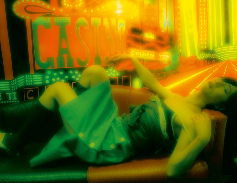An adult female reclines on a red couch in front of colorful neon casino signage in an indoor setting with low-key lighting.