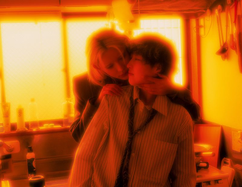 Woman in a blazer embraces a young adult in a striped shirt in a kitchen setting, bathed in warm, golden lighting.