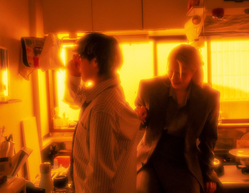 Indoor shot of two people in a kitchen; one in glasses and a striped shirt stands at the counter, while the other in a blazer sits.