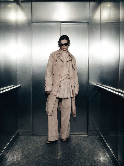 Adult female fashion model in a tan coat and pants stands in a metal elevator with a serious expression, illuminated by harsh light.