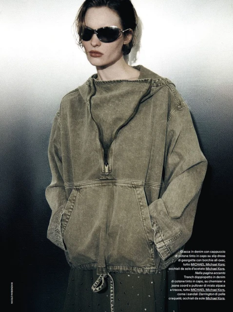 Fashion editorial shot: a slender woman in sunglasses and a hooded top poses against a muted background in a studio setting.