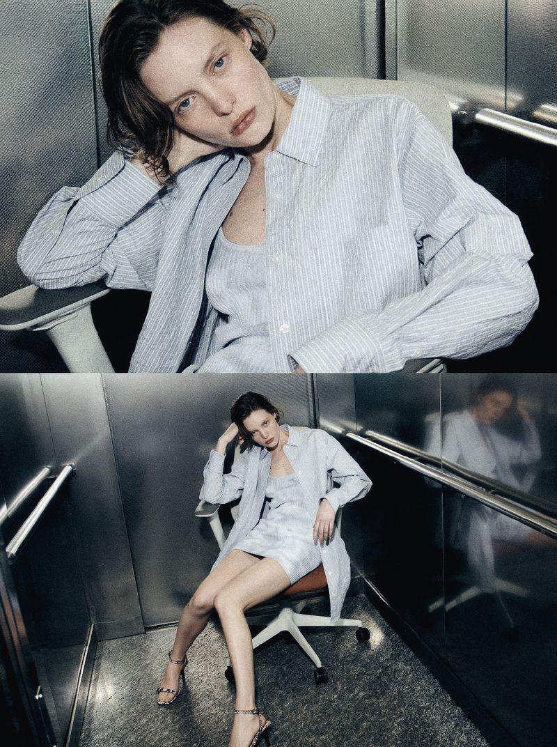 A young woman with brown hair sits in a rolling chair in an elevator wearing a striped blue shirt, a dress, and strappy heels.