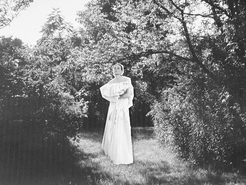 Monochrome portrait of a woman in an off-shoulder dress, posing in a natural setting with trees and foliage, creating a vintage aesthetic.