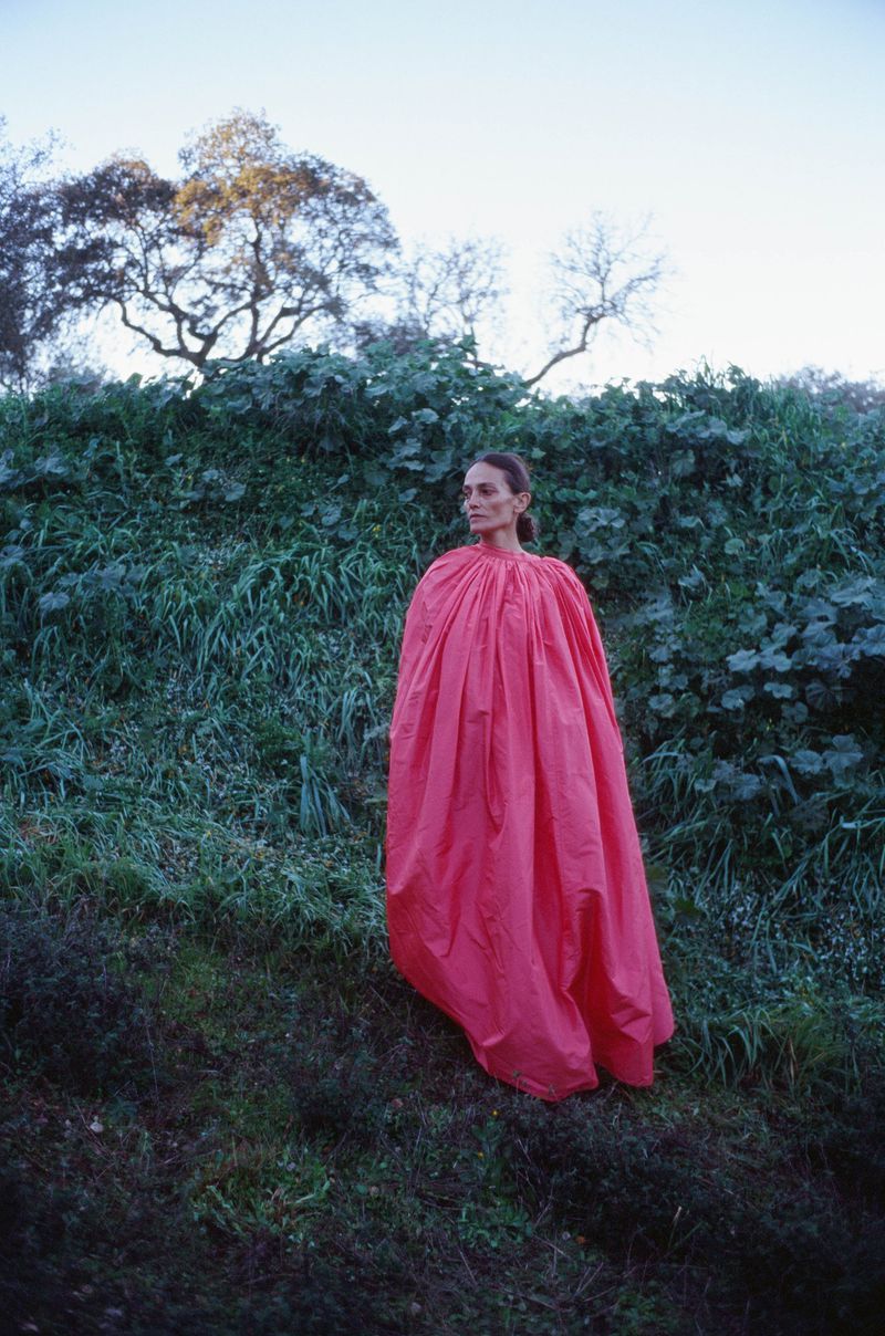 Woman in a red dress standing outdoors surrounded by green vegetation and trees under a light blue sky, creating a fashion or art image.