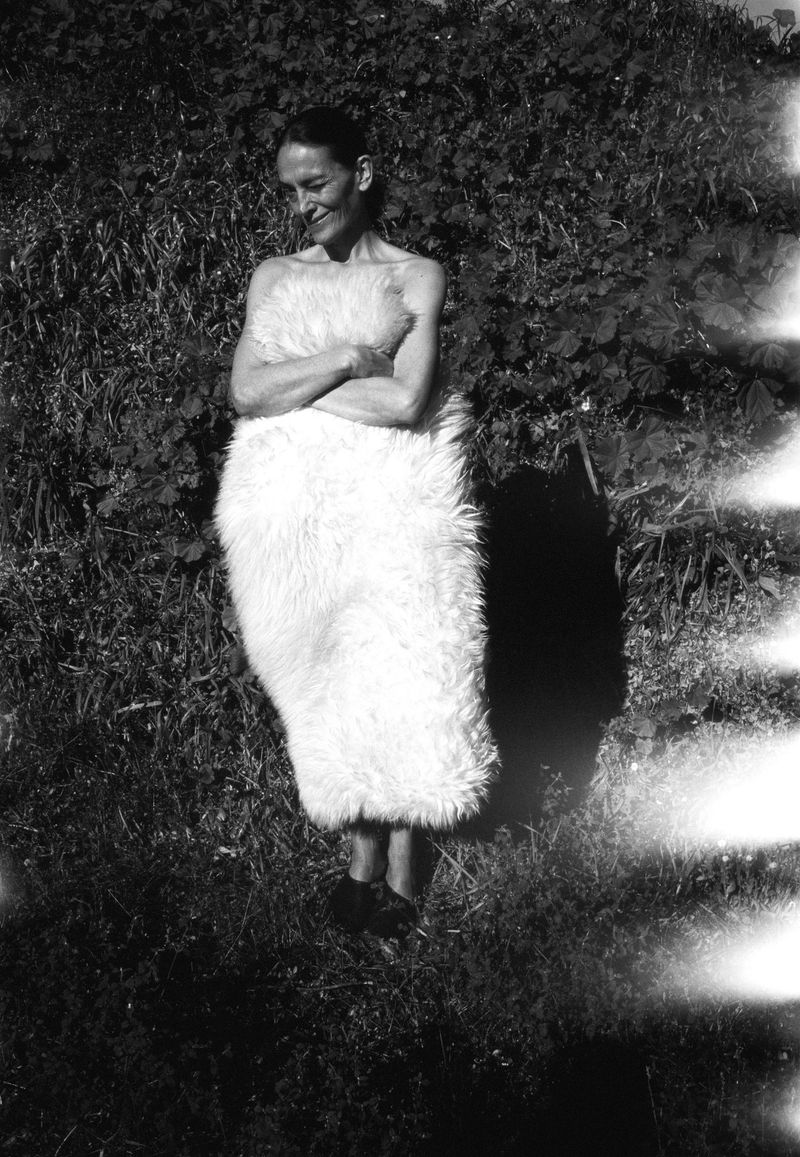Black and white shot of a woman in a fuzzy wrap standing outdoors surrounded by green foliage, captured with a film photography aesthetic.