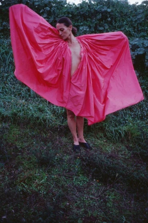 A woman poses in a pink garment amid lush greenery, gazing downward in a natural outdoor setting with soft lighting.