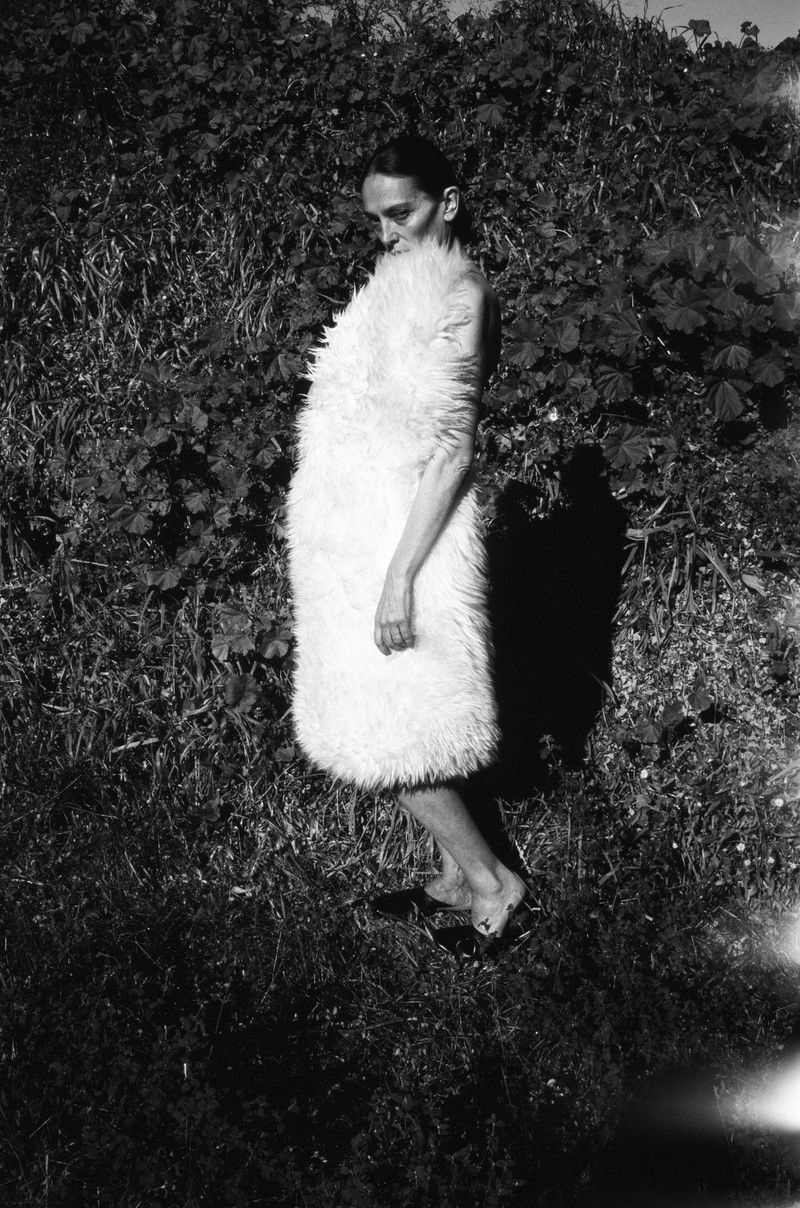 A woman in a light-colored, fluffy garment stands in a grassy outdoor setting in a black and white fashion photograph.