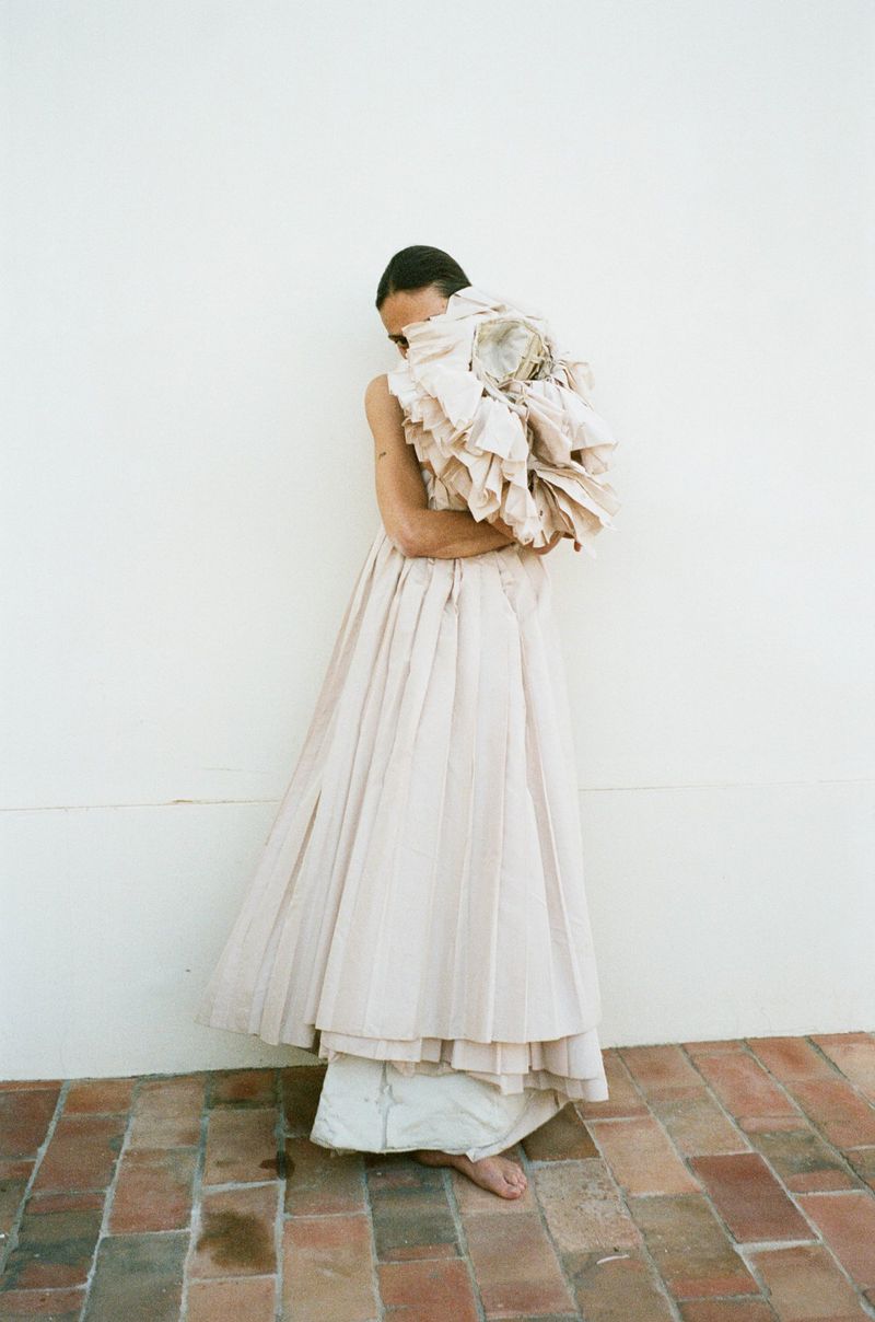 Person in long off-white dress stands barefoot against white wall, partially obscured by large ruffled fabric, creating an artistic fashion image.